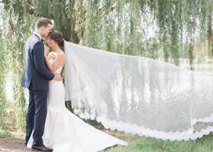 Groom kissing bride's forehead while holding each other in their arms outside in front of a weeping tree